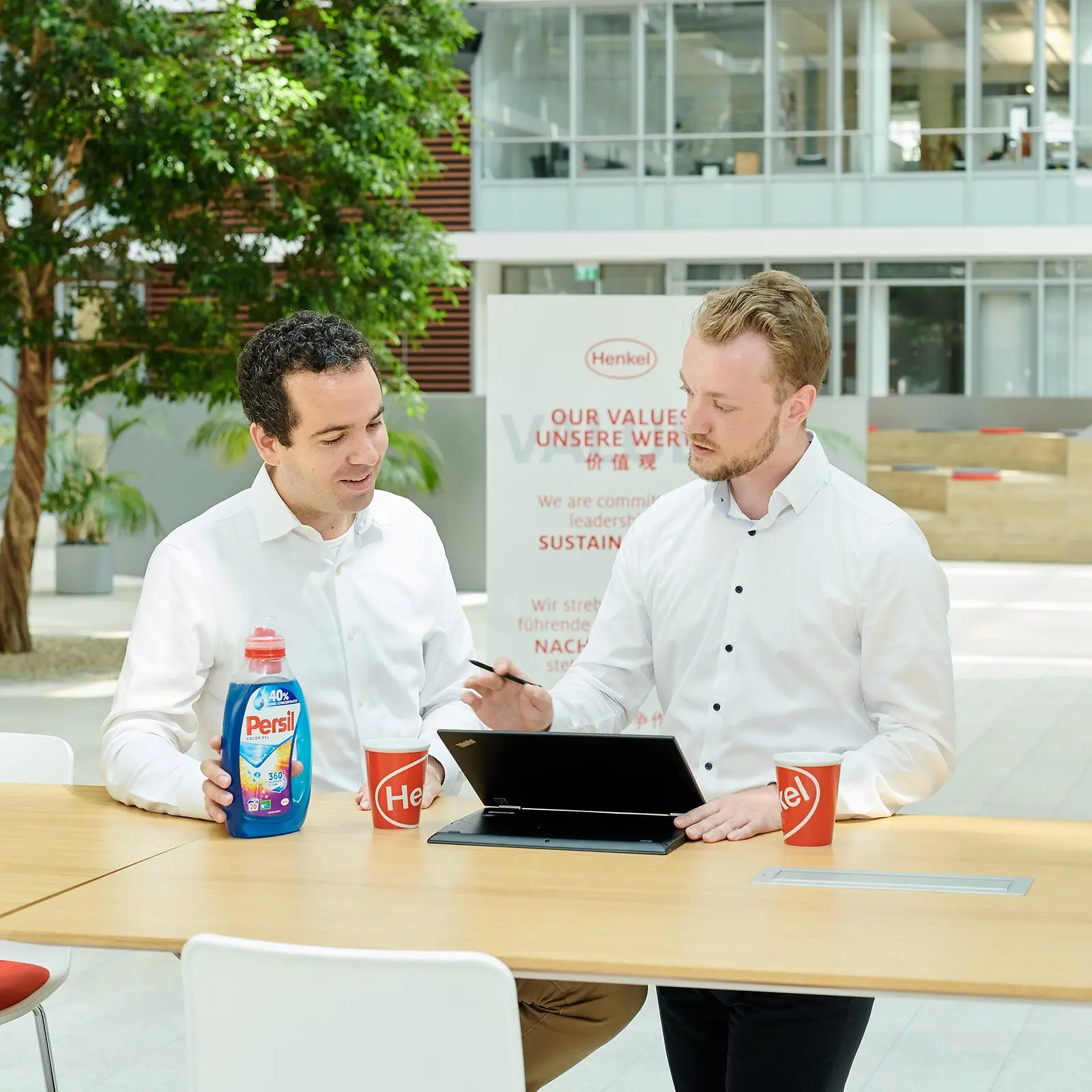 ผู้ชายสองคนยืนอยู่กับโต๊ะที่มีขวดผลิตภัณฑ์ Persilและแล็ปท็อปวางอยู่บนโต๊ะ