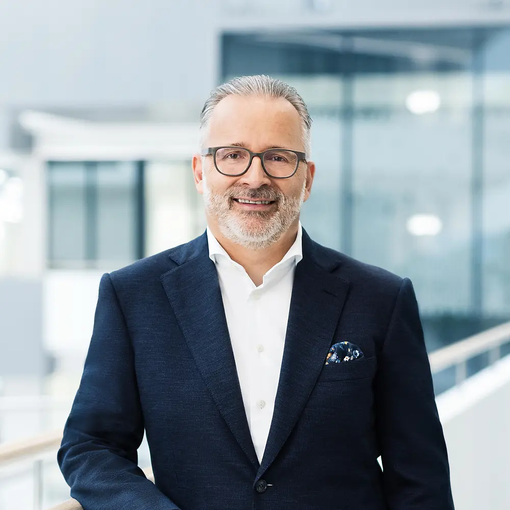 ภาพถ่ายผู้ชายที่สวมแว่นและเสื้อสูทสีดำโดยมีฉากหลังเป็นสำนักงาน