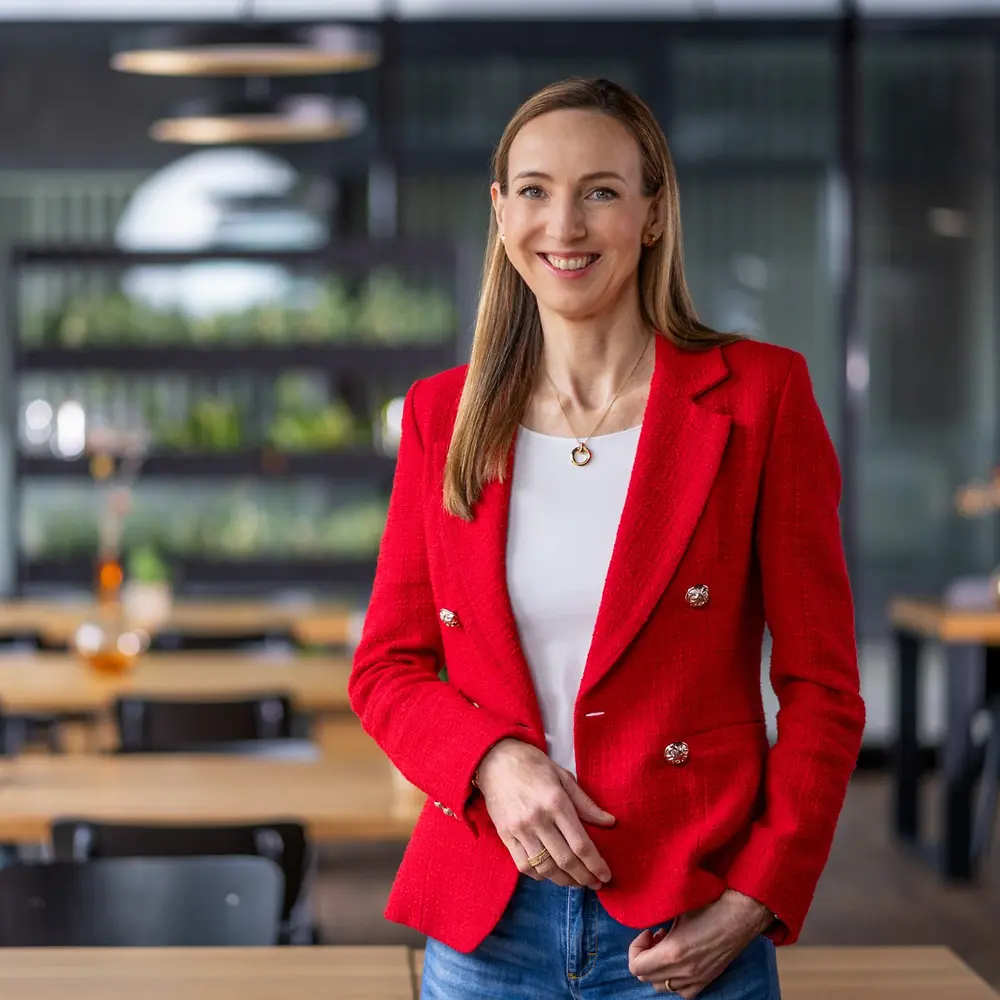 ดร. Simone Bagel-Trah สวม เบลเซอร์สีแดงสดและเสื้อสีขาว ยืนอยู่ในบรรยากาศแบบคาเฟ่สมัยใหม่ มีโต๊ะไม้และแจกันตกแต่งอยู่เบื้องหลัง